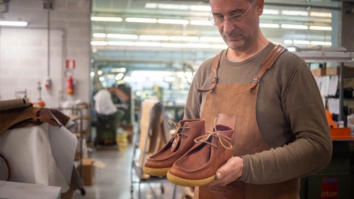 En skomakare iklädd läderförkläde håller ett par nytillverkade bruna Astorflex-skor i händerna.