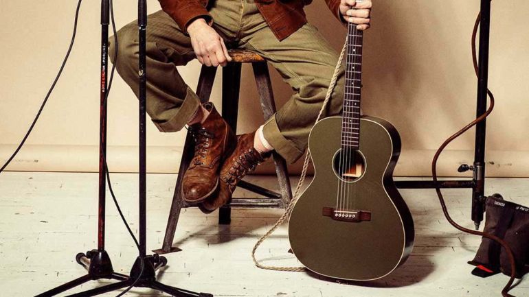 Red Wing: A musician seated on a high wooden stool wearing beautifully worn Red Wing boot and holding an acoustic guitar.