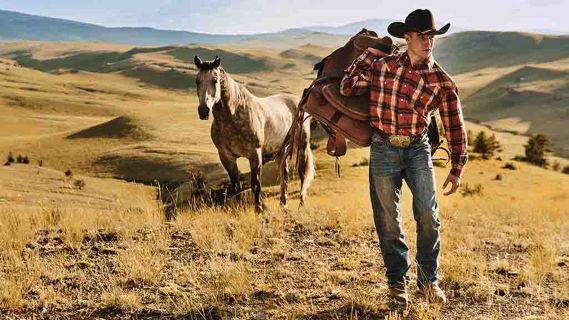 En robust cowboy, klädd i traditionell klädsel inklusive en Stetson-hatt, promenerar tillsammans med en majestätisk häst mitt i det hisnande bergslandskapet.